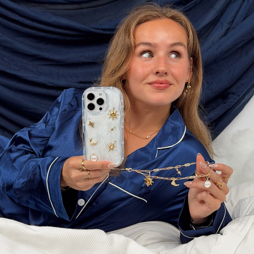 Woman wearing satin navy pyjamas holding a phone in a stardust after dark phone case with gold charm embellishments. The phone has a matching gold chain phone strap with celestial style charms, including a moon, sun, star, diamond shape and 11:11 charm. 