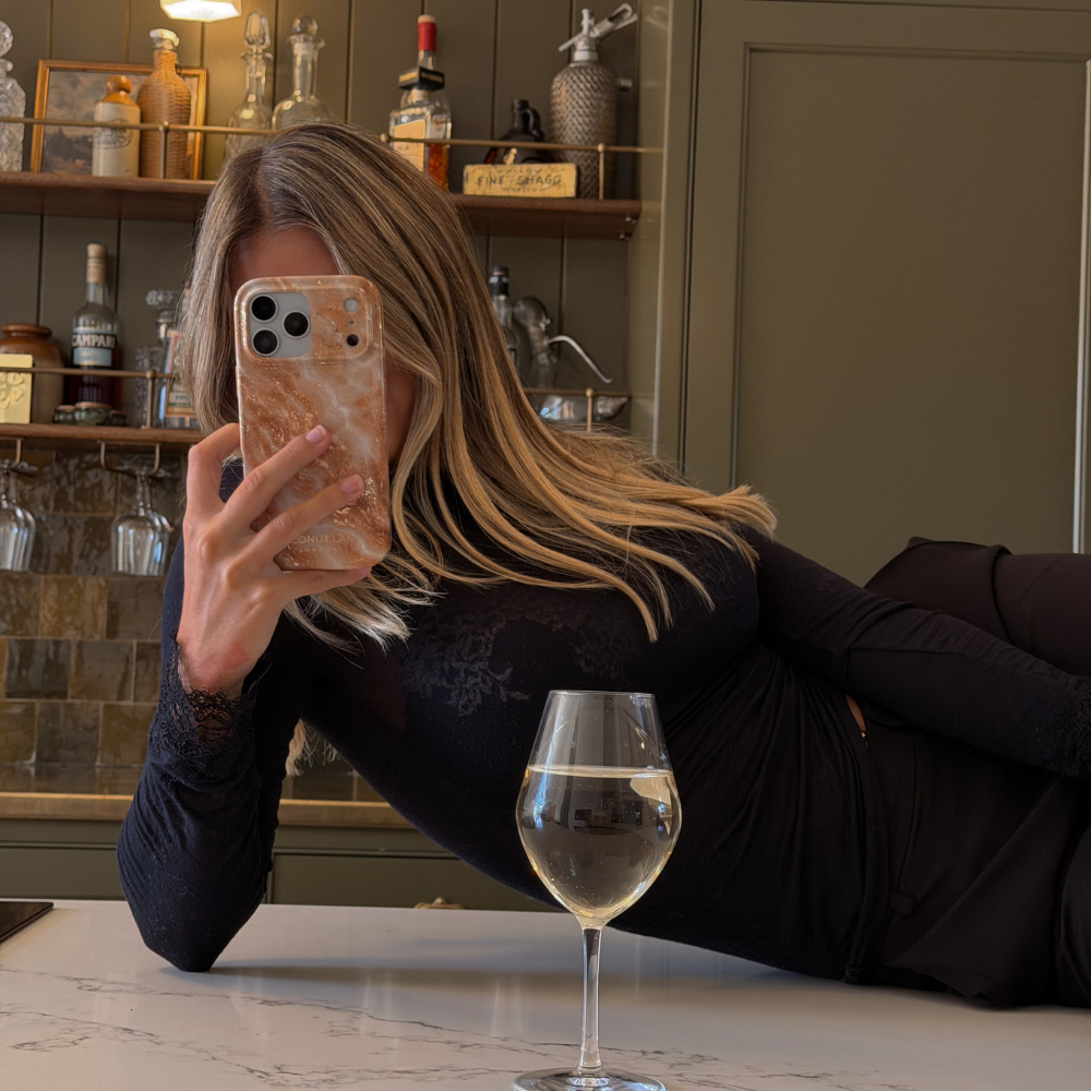 Woman with blonde hair wearing a black long sleeve top is holding an iphone in a marble effect phone case with cream, gold, light brown and white tones running through it.