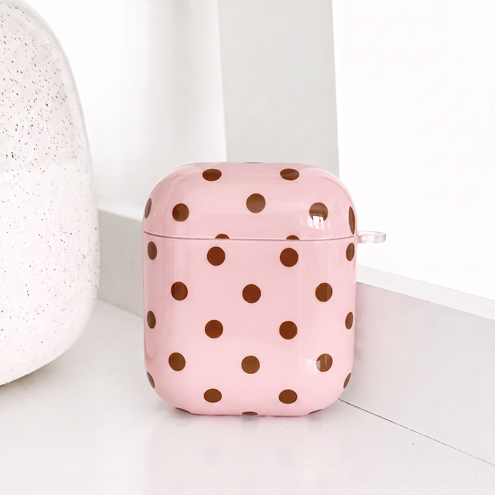 A glossy pink case for AirPods with brown polka dots and a small clear clip loop on the side. It sits on a white shelf beside a white speckled vase.