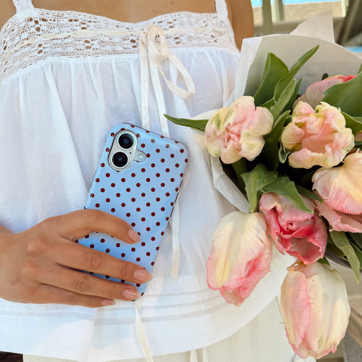 Girl holding her brown and blue polka dot phone case in hand wearing a spring skirt and pink flowers in hand