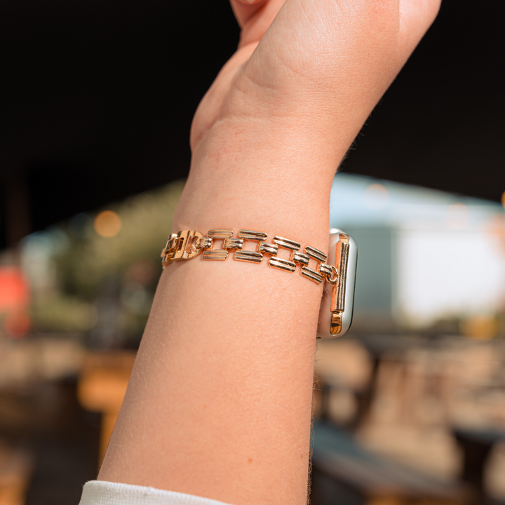 A close-up shot of a light-skinned wrist showcasing a **Square Gold Bracelet Apple Watch Strap** by **Coconut Lane**. The rectangular watch face of the Apple Watch is partially visible, while the background is artistically blurred, capturing indistinct outdoor elements.