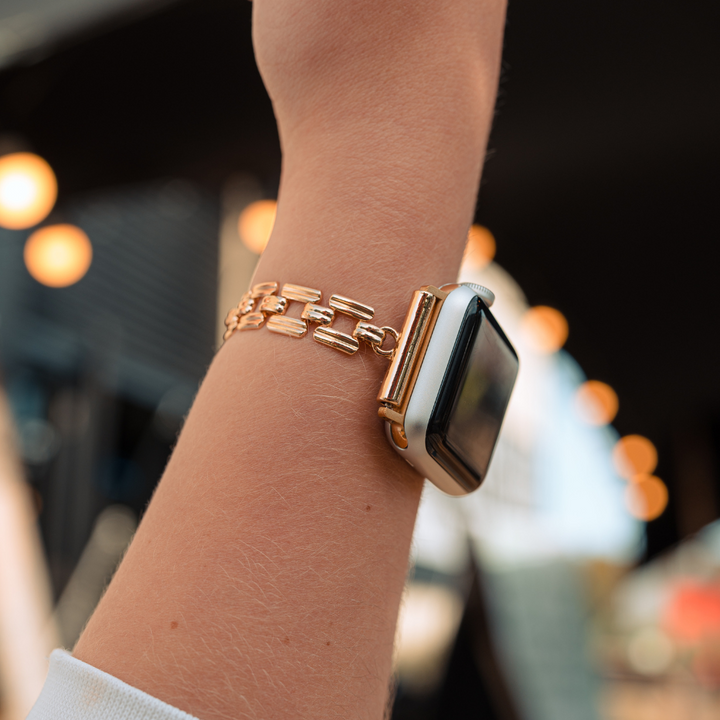 A person wearing a Coconut Lane Square Gold Bracelet Apple Watch Strap. The watch screen is black, possibly turned off, and the photo is taken from the side, focusing on the watch and wrist. The background is blurred with warm lights and distant structures.