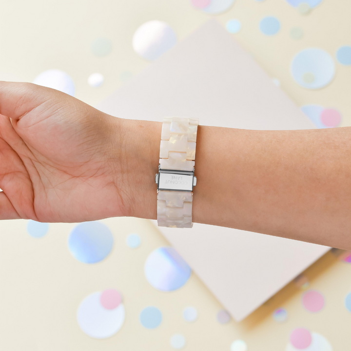 A hand wearing a Luxe Iridescent Apple Watch Strap by Coconut Lane, featuring an iridescent finish and a silver clasp. The background is adorned with pastel-colored circles on a soft, light pink platform.