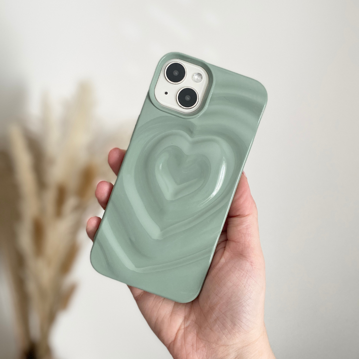 A hand holding a light green smartphone with Coconut Lane's Melting Heart Phone Case in Sage, featuring an extra protective case with a raised heart design. The background is blurred with neutral tones.