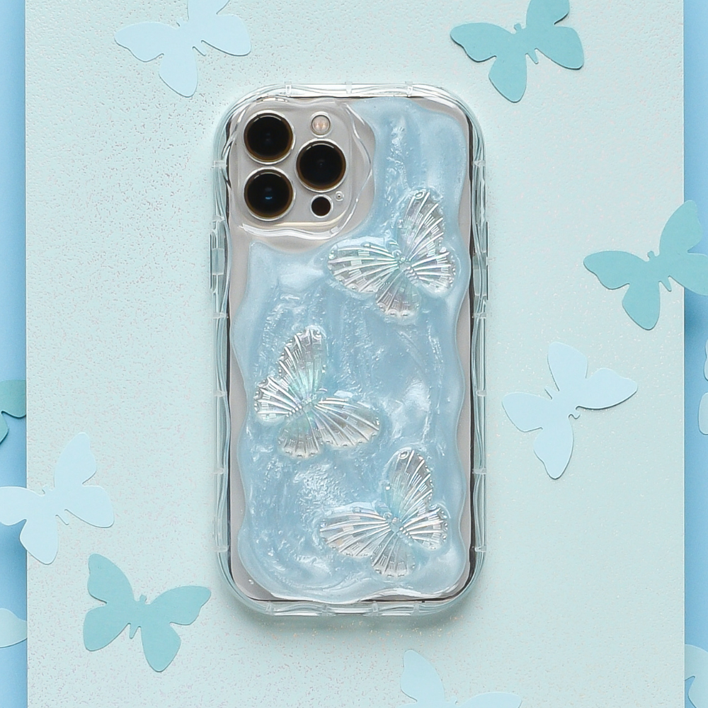 A smartphone encased in the Shimmer Blue Butterflies Phone Case by Coconut Lane, which is clear and extra protective with a wavy texture resembling flowing water, lies on a light blue background adorned with scattered paper butterfly cutouts. The phone has a triple-lens camera system.