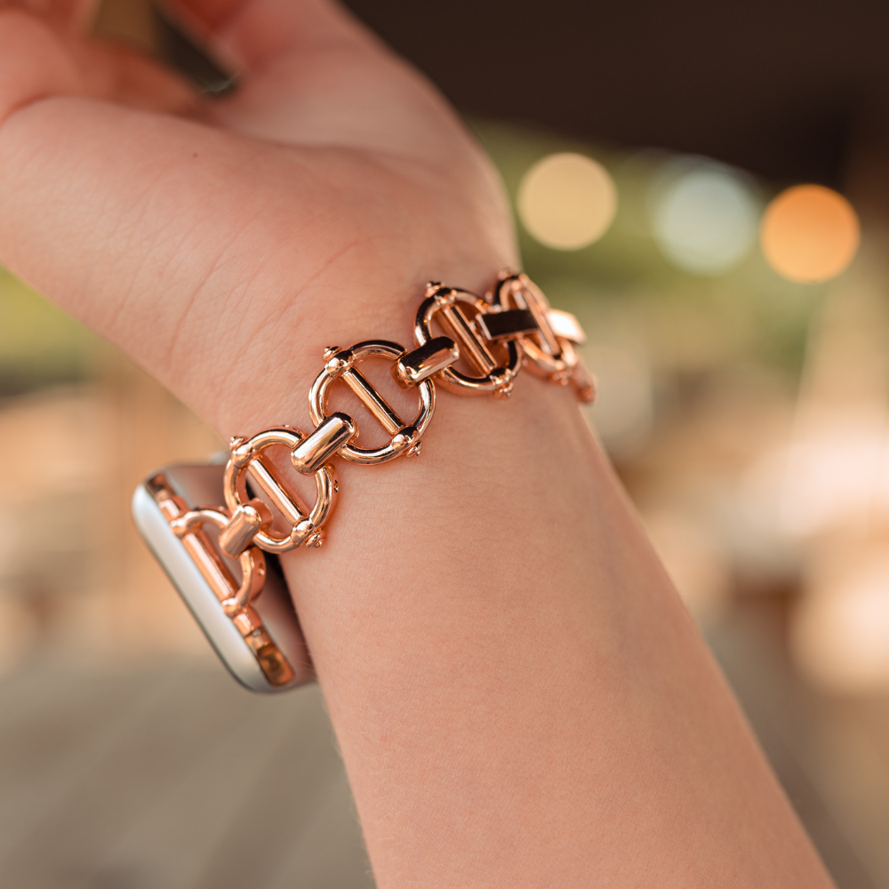 Close-up of a person's wrist wearing the Metal Link Apple Watch Strap - Rose Gold by Coconut Lane. The background is softly blurred with warm tones, suggesting a cozy, indoor setting. This stainless steel strap features interlocking links, contributing to its elegant and stylish appearance.