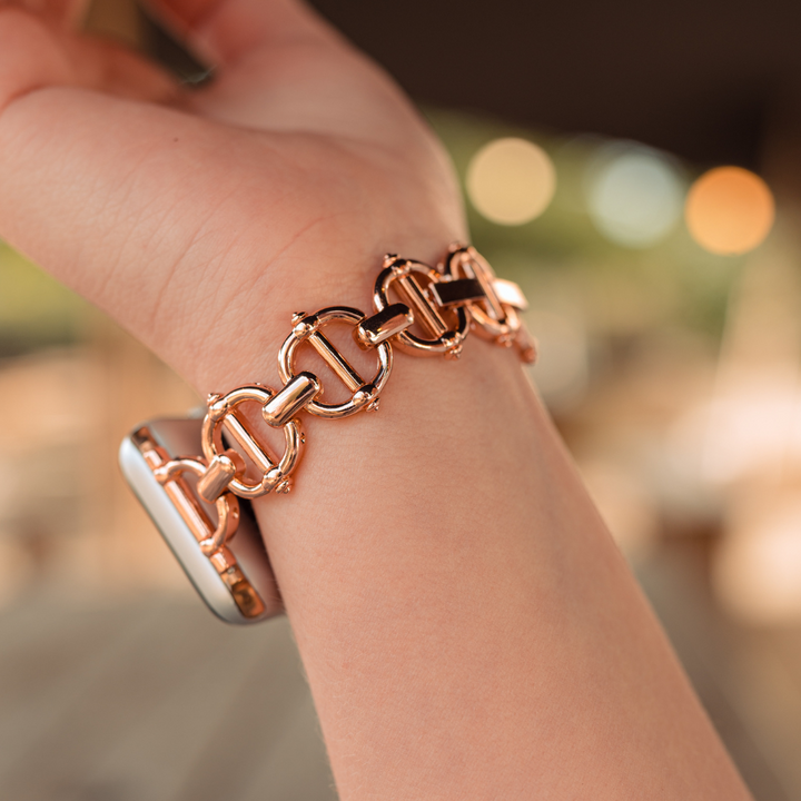 Close-up of a person's wrist wearing the Metal Link Apple Watch Strap - Rose Gold by Coconut Lane. The background is softly blurred with warm tones, suggesting a cozy, indoor setting. This stainless steel strap features interlocking links, contributing to its elegant and stylish appearance.