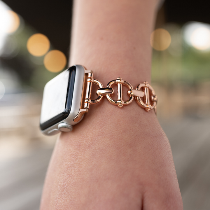 A close-up image of a person's wrist wearing an Apple Watch adorned with the Coconut Lane Metal Link Apple Watch Strap in Rose Gold. The background is softly blurred with warm lights, giving the scene a stylish and sophisticated ambiance.