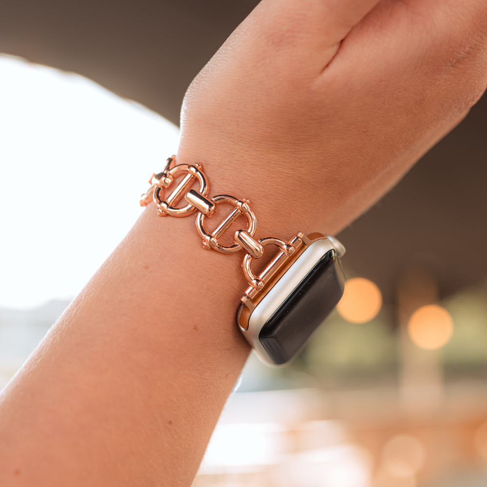 A close-up of a woman's wrist showcases the Coconut Lane Metal Link Apple Watch Strap in Rose Gold, elegantly attached to a modern smartwatch. The blurred background accentuates the sophisticated blend of traditional jewelry design with contemporary technology.
