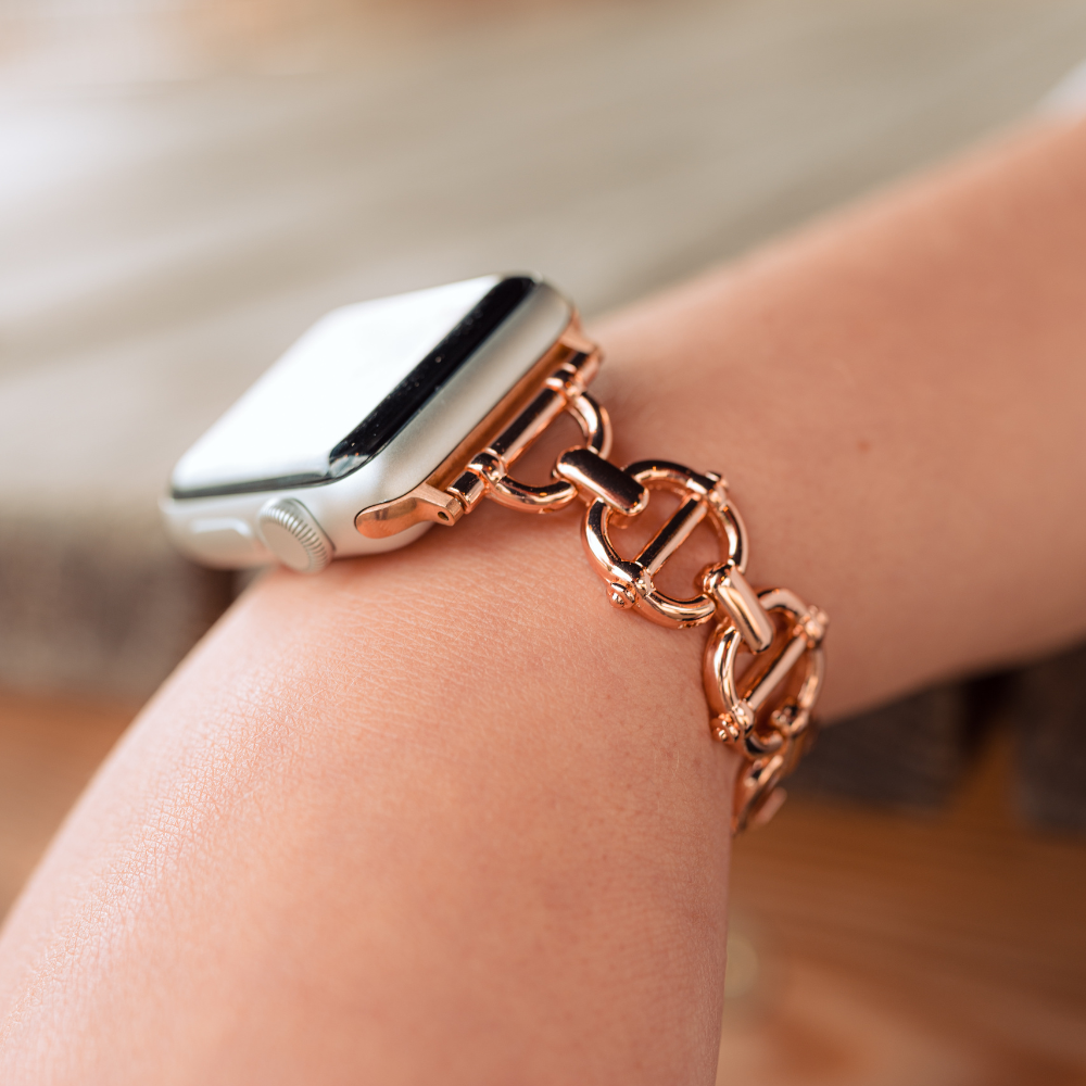 A close-up image shows a person's wrist adorned with an Apple Watch featuring the exquisite Metal Link Apple Watch Strap in rose gold from Coconut Lane. The stainless steel band, finished in elegant rose gold, enhances the sophistication of the tech accessory. In this shot, the background is blurred to keep all attention on the watch and its stunning strap.