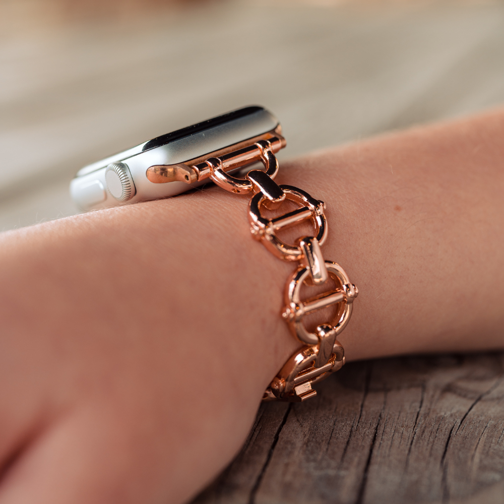 A close-up of a wrist wearing a Metal Link Apple Watch Strap - Rose Gold by Coconut Lane. The background is blurred but features a wooden surface. This sleek strap adds an elegant touch to any outfit.