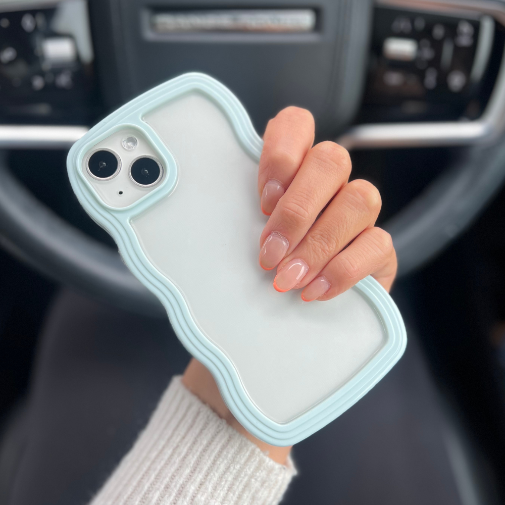 A person's hand is holding up a white iPhone15 encased in a light blue, protective Curvy Phone Case by Coconut Lane. The case highlights the back of the phone with its dual camera lenses. In the background, you can see the interior of a car, including the steering wheel and dashboard. The person's nails are neatly manicured with a natural look.