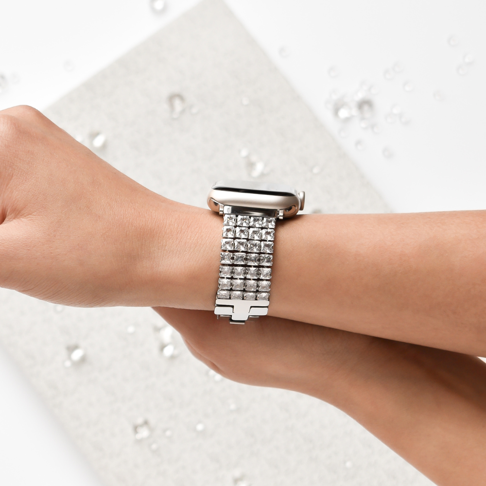 A person wearing the Coconut Lane Sparkle Super Glam Apple Watch Strap, adorned with stainless steel and encrusted with gemstones. The wearable device, featuring an adjustable strap, is showcased prominently on their wrist, set against a light textured background with scattered sparkling gems.