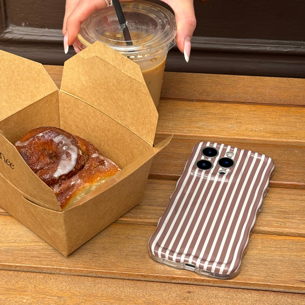 Hand holding iced coffee, cinnamon roll, and phone with Coconut Lane chestnut striped wavy phone case on a table.