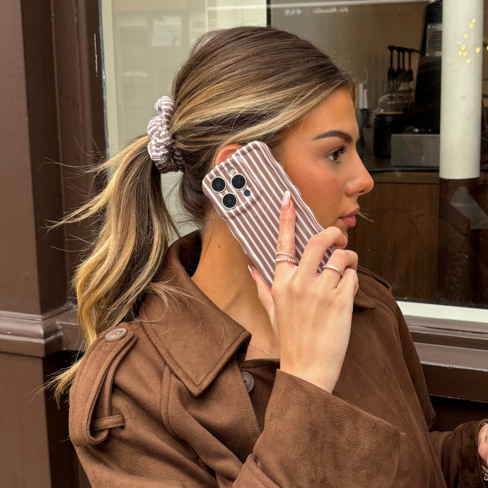 Blonde woman with ponytail holds a Coconut Lane phone case with chestnut wavy stripes to her ear outside a café.