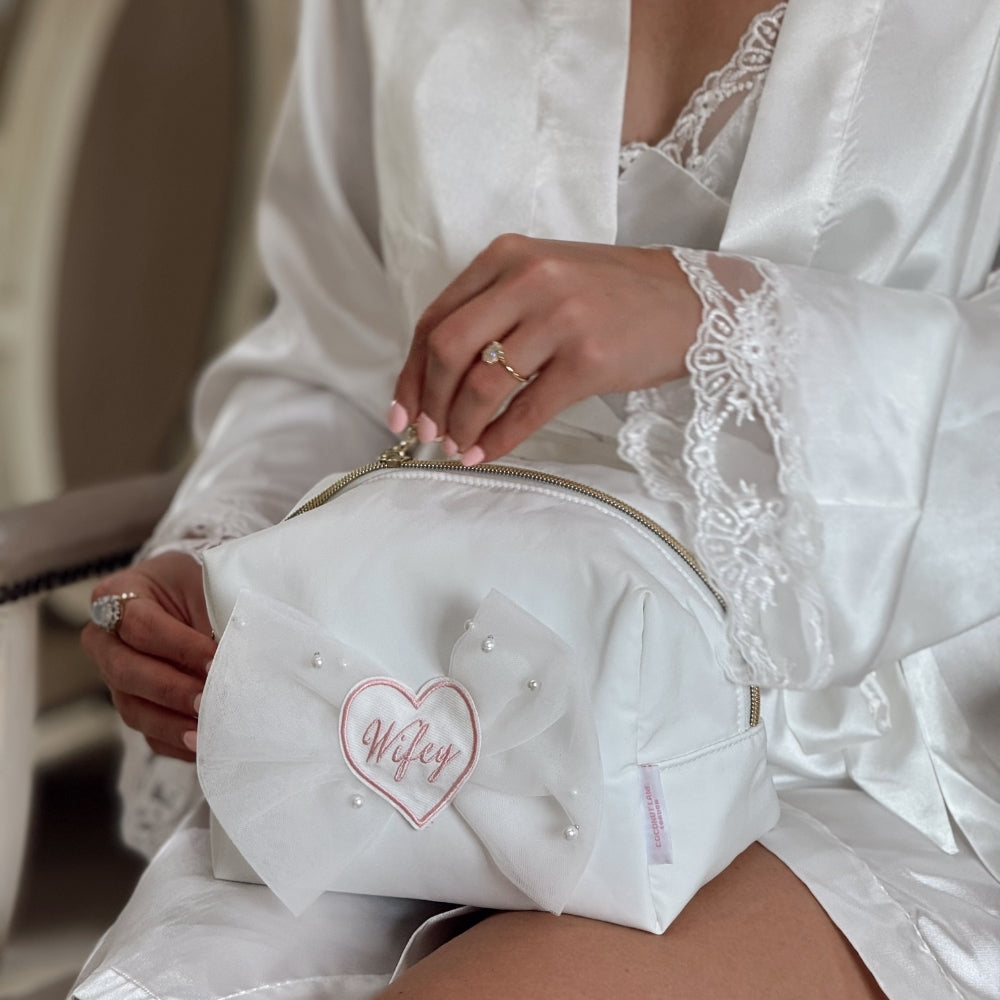 Bride holding a makeup bag with 'Wifey' embroidered on it, wearing a white satin robe.