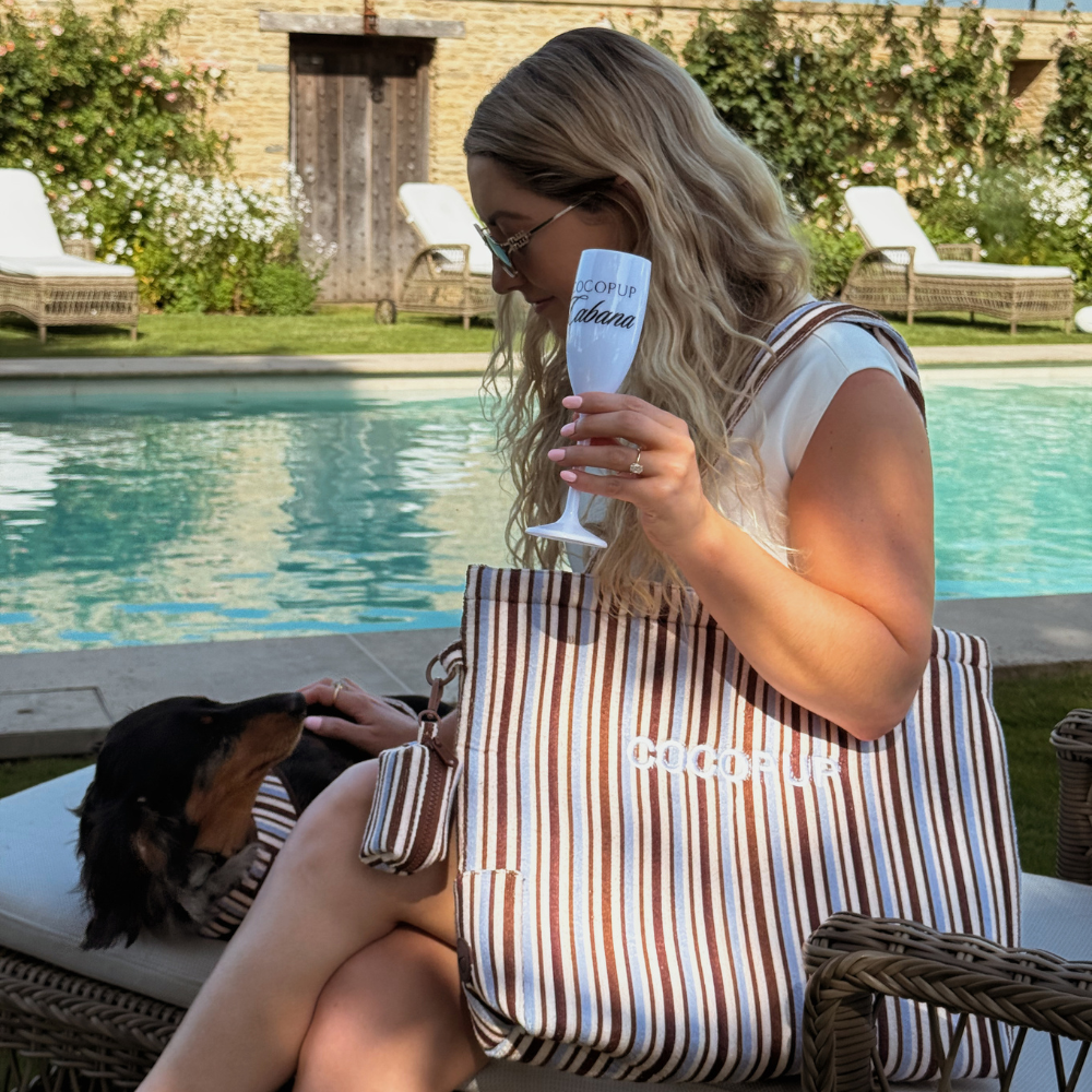 Woman with wavy blonde hair by a pool holds a drink, her small dog peeking from a brown and blue striped tote bag.