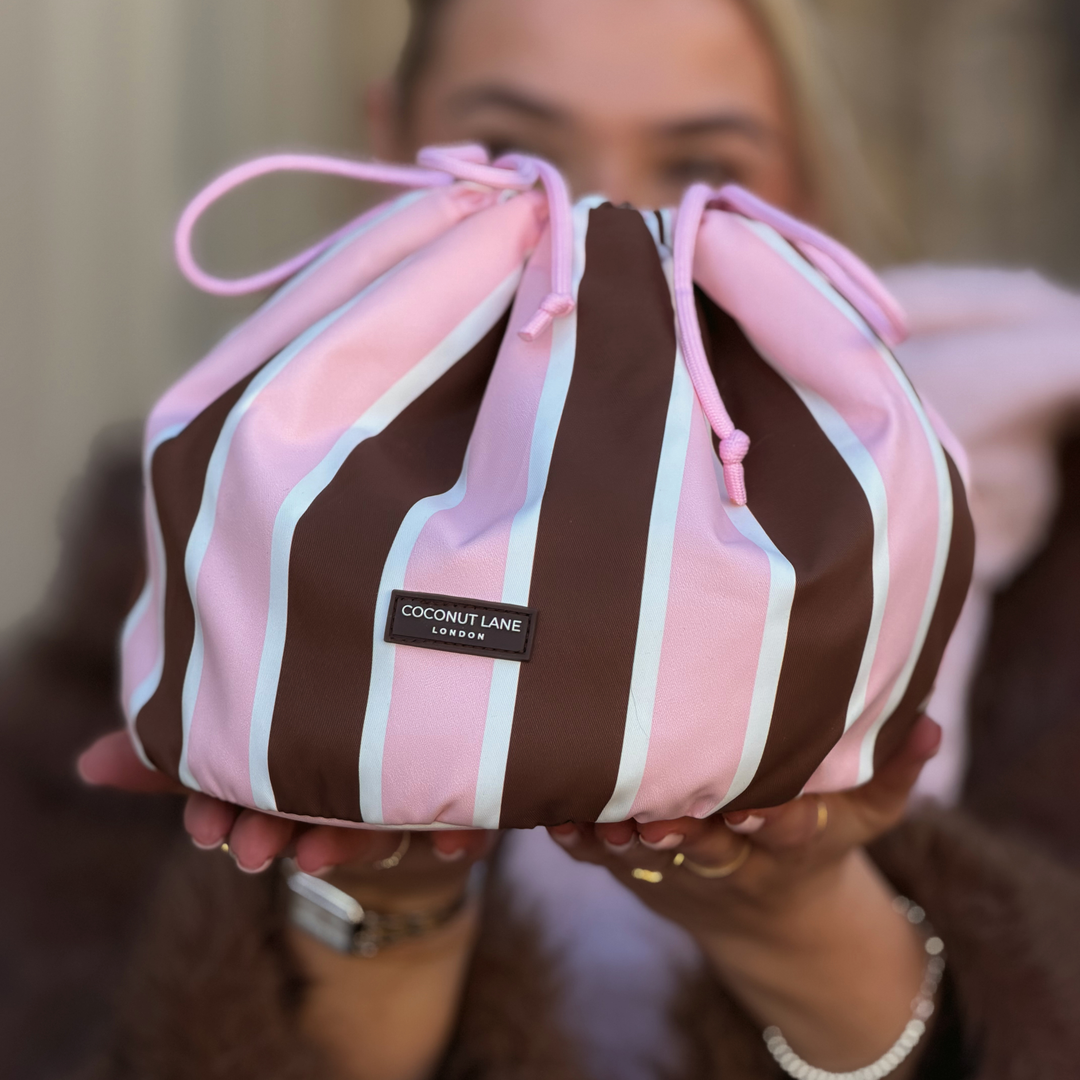 Woman wearing a baby pink scarf and brown faux fur jacket, holding a drawstring toiletry bag featuring chocolate brown, baby pink and white stripes with pink drawstring closure.