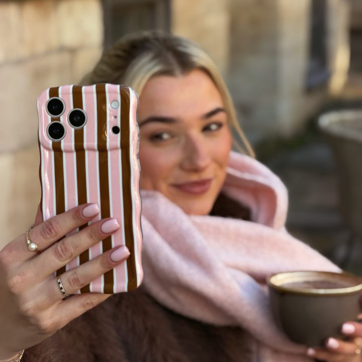 woman wearing baby pink scarf and brown faux fur coat holding an iphone in a cocoa & Pink Stripe Wavy Phone Case featuring glossy brown, blush pink and cream vertical stripes, photographed diagonally on a pink background