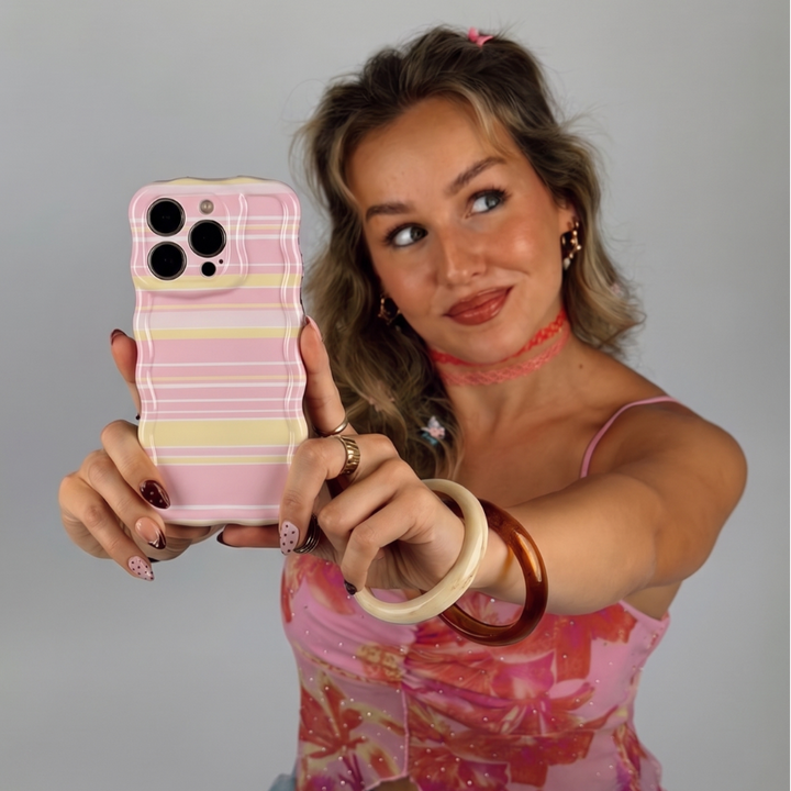 A woman taking a selfie with a phone case featuring a pastel pink, yellow, and white wavy stripe pattern. She is wearing a pink patterned top and various bracelets.