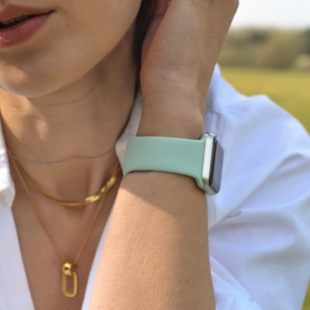 Close-up of a person sporting a Coconut Lane NAKD Apple Watch Strap in Cool Mint on their wrist. They wear a white shirt, a gold chain, and a pendant necklace. The background captures green outdoor scenery with an out-of-focus horizon, but the focus remains on the wrist and part of the face.