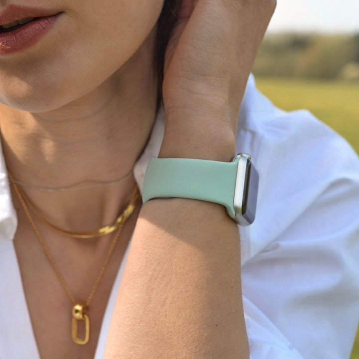 Close-up of a person sporting a Coconut Lane NAKD Apple Watch Strap in Cool Mint on their wrist. They wear a white shirt, a gold chain, and a pendant necklace. The background captures green outdoor scenery with an out-of-focus horizon, but the focus remains on the wrist and part of the face.