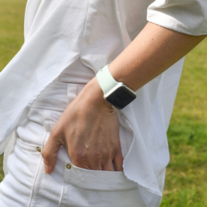 A person wearing a white shirt and light-colored pants stands with their hand in their pocket. They are sporting an Apple Watch with a Coconut Lane NAKD Apple Watch Strap - Cool Mint on their wrist. The background is an out-of-focus green grass area.