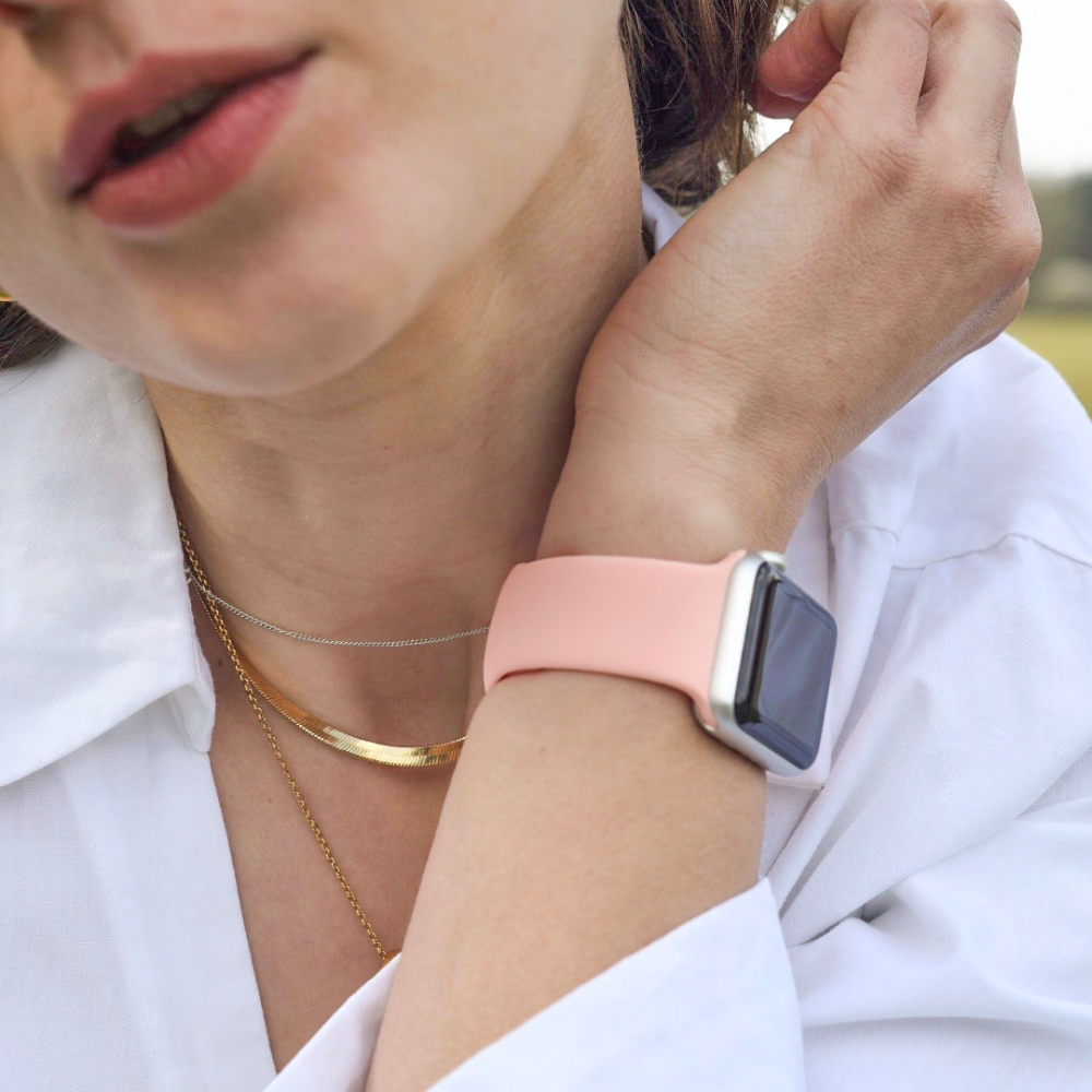 A close-up image of a person with light skin wearing a white shirt, a gold necklace, and an Apple Watch with the Coral Pink NAKD strap from Coconut Lane. The person is touching their face with one hand, showcasing the durable silicone strap prominently displayed on their wrist, which blends perfectly with spring-inspired pastels.
