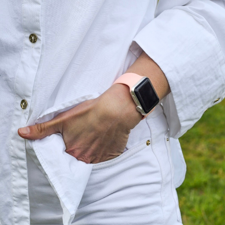 A person wearing a white long-sleeve shirt and white pants with gold buttons stands with one hand in their pocket, sporting the Coral Pink NAKD Apple Watch Strap by Coconut Lane on their wrist. The backdrop of green grass adds a spring-inspired pastel vibe to the scene.