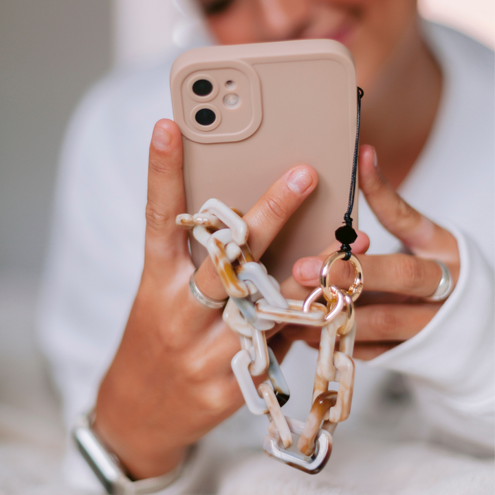 A person is holding a beige smartphone with a chunky plastic chain attached to the Coconut Lane NAKD Phone Case - Nude. They are wearing multiple rings on their fingers and appear to be using the phone, smiling. The background is soft and slightly blurred, highlighting the case's full camera protection.