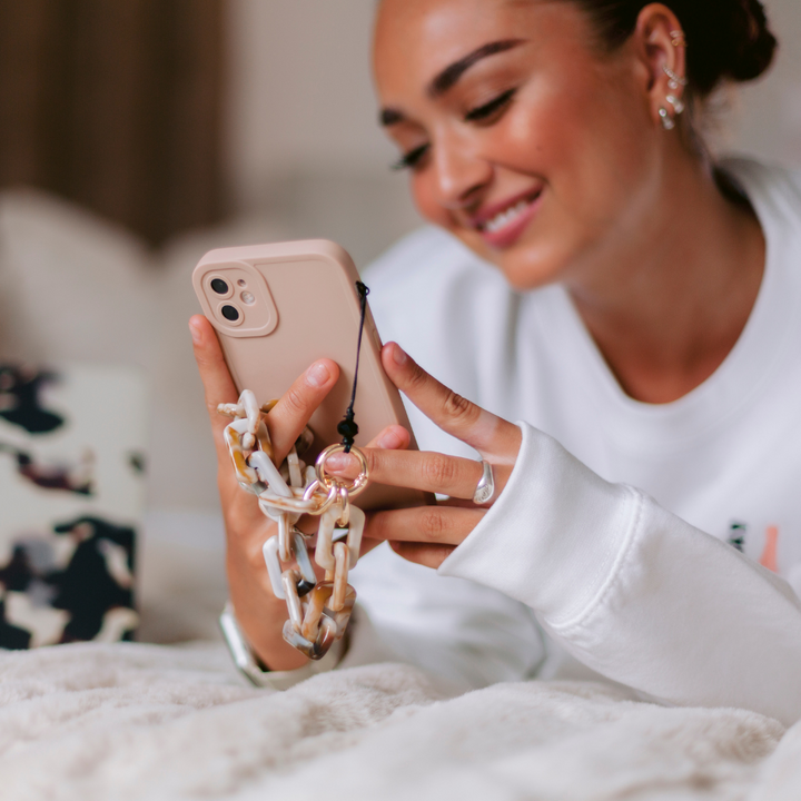 A woman dressed in a white sweater is lying on a bed, smiling as she looks at her pink smartphone with her Coconut Lane NAKD Phone Case - Nude. She has several earrings and a ring on her finger, holding the phone with a chain strap attached. A laptop with a camouflage cover is partially visible in the background.