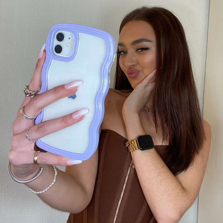 A young woman with straight, dark hair is taking a selfie using her iPhone15, which is protected by the highly durable Curvy Phone Case - Lilac from Coconut Lane. She is dressed in a brown top and accessorized with several rings, bracelets, and a watch. Her lips are pursed in a playful facial expression.