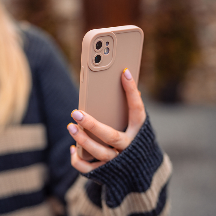 A person with light purple nail polish holds a beige NAKD Phone Case - Nude from Coconut Lane, ensuring full camera protection. They are wearing a dark blue sweater with beige stripes. The soft, flexible TPU case complements the cozy, blurred background featuring some greenery.