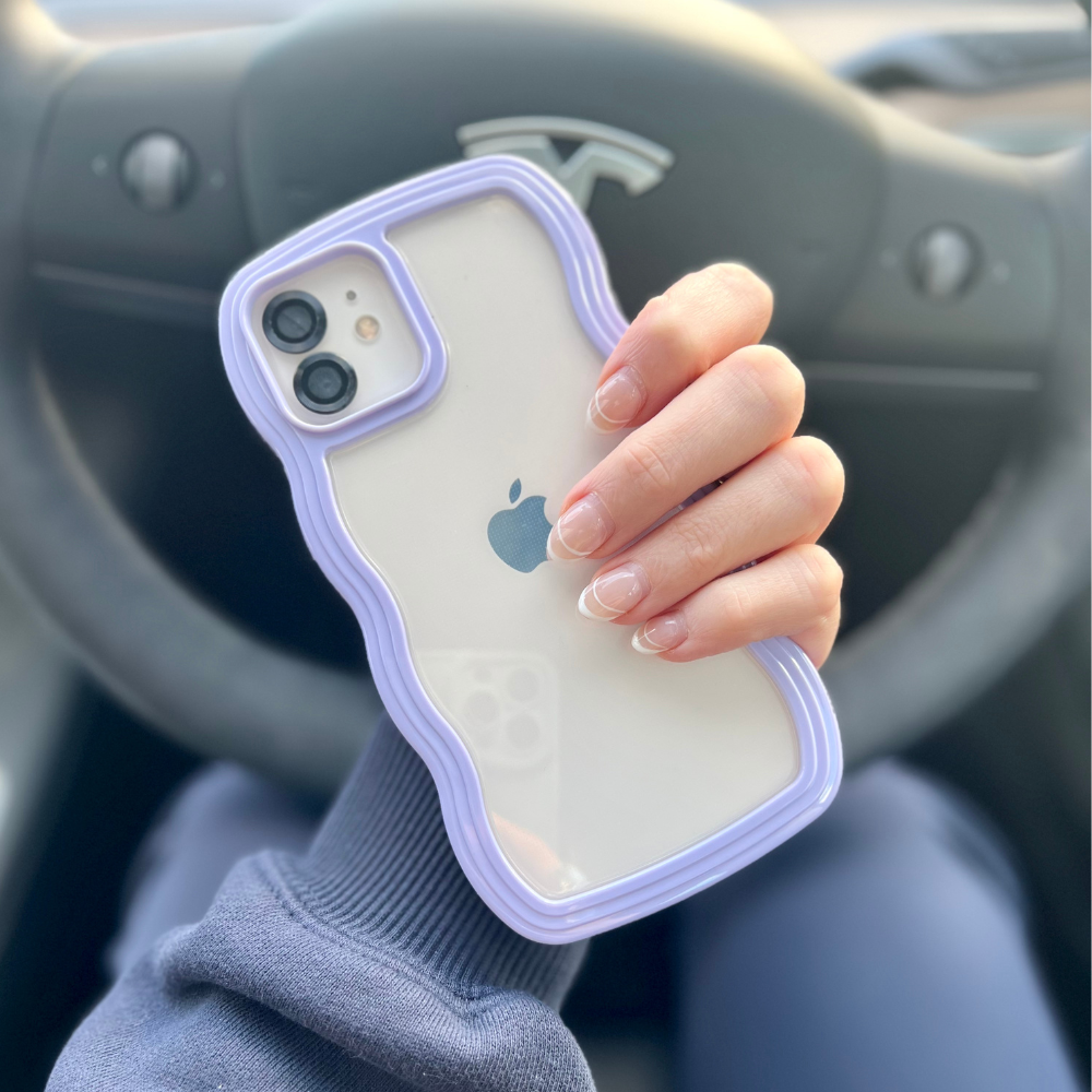 A hand with manicured nails holds a white iPhone 15 encased in Coconut Lane's highly protective Curvy Phone Case in lilac, in front of a car's steering wheel bearing a "T" shape logo. The person appears to be wearing a long-sleeve shirt.