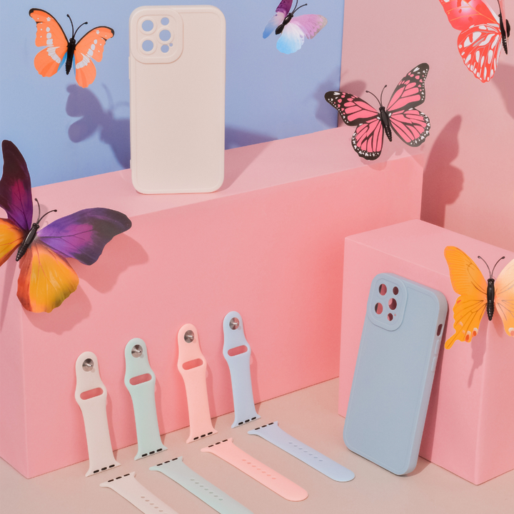A display showcasing Coconut Lane's NAKD Collection of phone cases and smartwatch bands in pastel colors. The scene features a light blue phone case and a light pink one, each with matching silicone Apple Watch straps in shades including the NAKD Apple Watch Strap - Cool Mint. The background is adorned with colorful, vibrant butterflies.