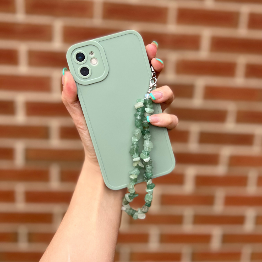 A hand with painted nails holds the NAKD Phone Case - Sage from Coconut Lane, encasing a green phone in its soft, flexible TPU cover. Attached to the case is a wrist strap made of green stone beads, ensuring full camera protection. A blurred brick wall serves as the background.