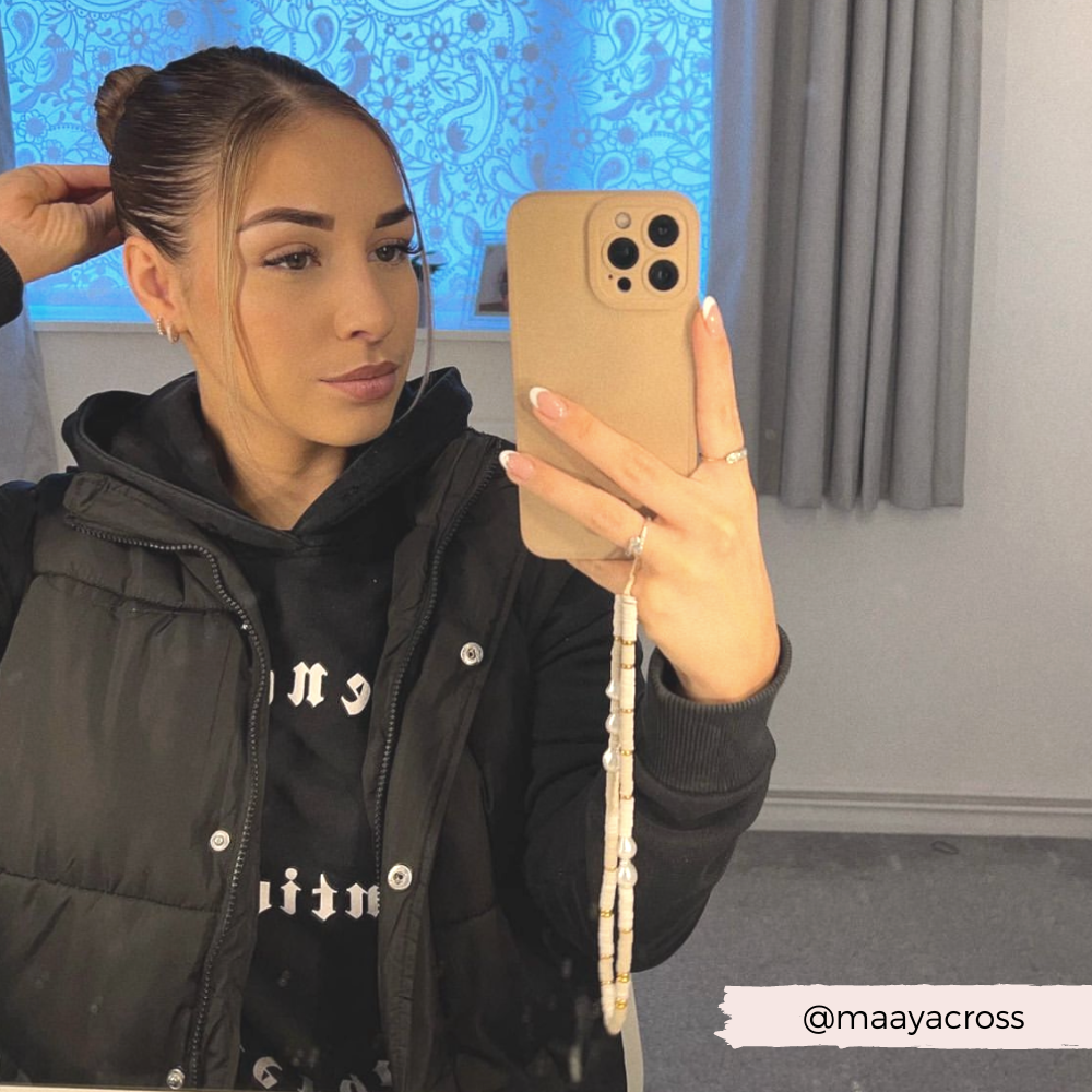 A woman with a bun hairstyle and light makeup stands in a room adorned with light blue patterned curtains and grey drapes, taking a mirror selfie. She is wearing a black hoodie and puffer vest, while holding her phone protected by the Coconut Lane NAKD Phone Case - Nude, which features full camera protection and is made of soft flexible TPU. Her Instagram handle is @maayacross.
