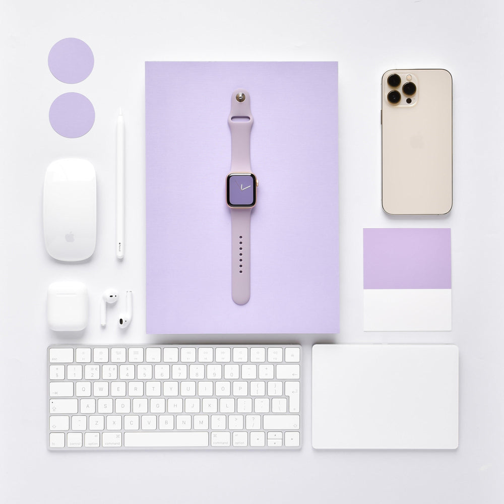 A neatly arranged flat lay of tech gadgets and accessories, including a lavender smartwatch with the NAKD Apple Watch Strap - Lavender Lilac from Coconut Lane, a silver smartphone, a wireless mouse, a stylus, earbuds, a keyboard, and a touchpad, all on a white and lavender background.