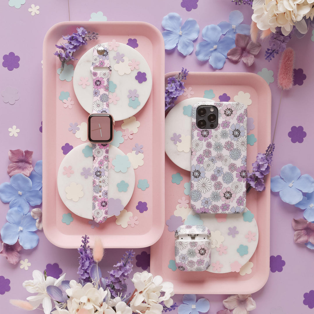 A pastel-themed flat lay showcases a smartwatch adorned with the Coconut Lane Pastel Flowers Apple Watch Strap, a smartphone with matching phone cases, and coordinating wireless earbuds. All items are elegantly arranged on pink trays, surrounded by pastel flowers and petals on a light purple background.