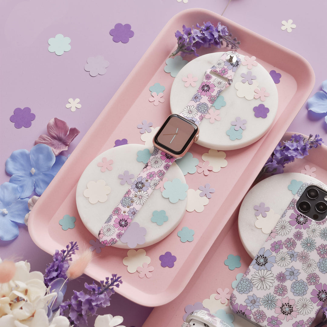 A smartwatch adorned with Coconut Lane's Pastel Flowers Apple Watch Strap sits on a pink tray, encircled by vibrant paper flower cutouts. Delicate purple flowers are arranged nearby, complementing a phone case decorated with a matching floral print. The scene is set against a backdrop of soft pastel lilac and pink shades.