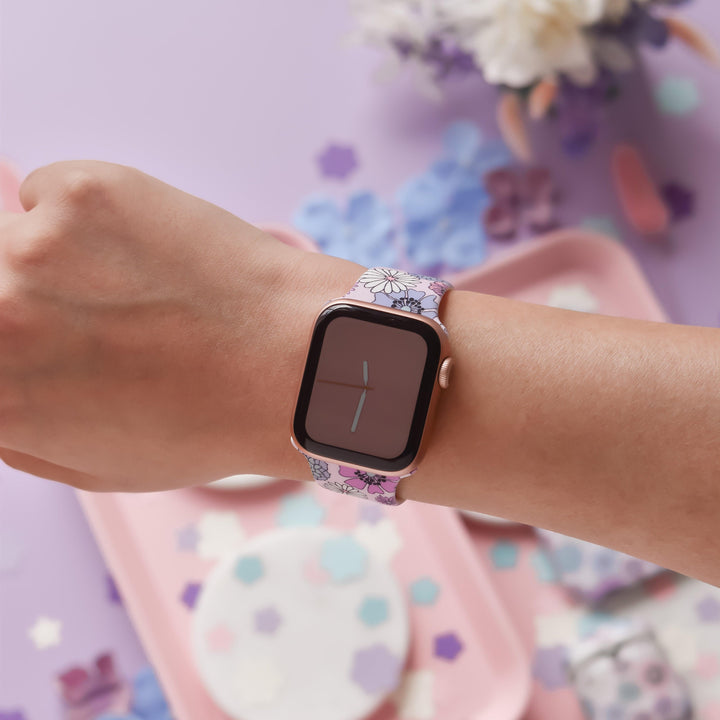 A person's wrist adorned with Coconut Lane's Pastel Flowers Apple Watch Strap. The watch face displays the time with minimalistic clock hands. The background includes pastel-colored decorations and shapes, creating a soft and colorful atmosphere that perfectly complements matching phone cases.