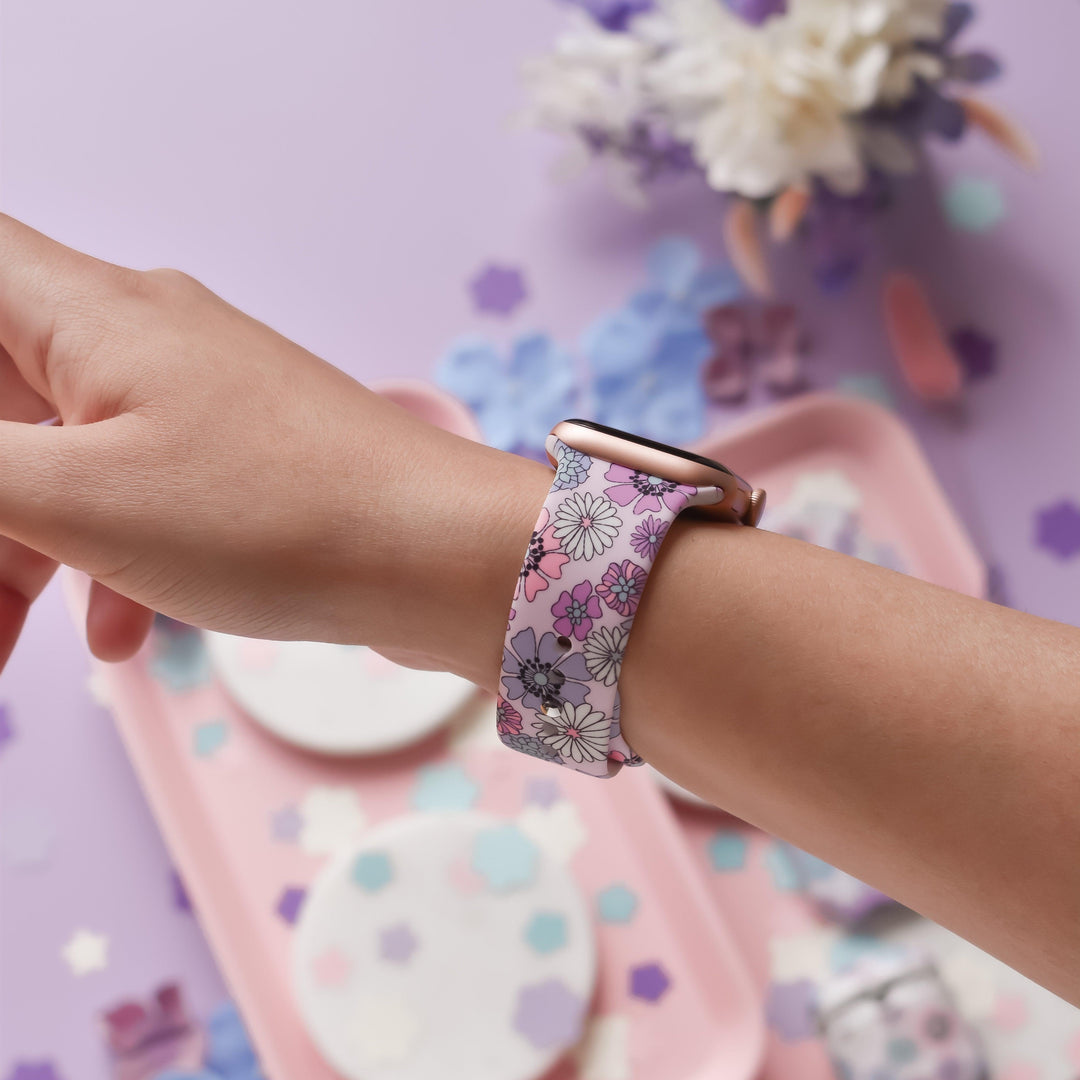 A person wearing a smartwatch with the Coconut Lane Pastel Flowers Apple Watch Strap on their left wrist. The background is decorated in pastel pink and purple with various objects and flower cutouts, creating a colorful and crafty setting.
