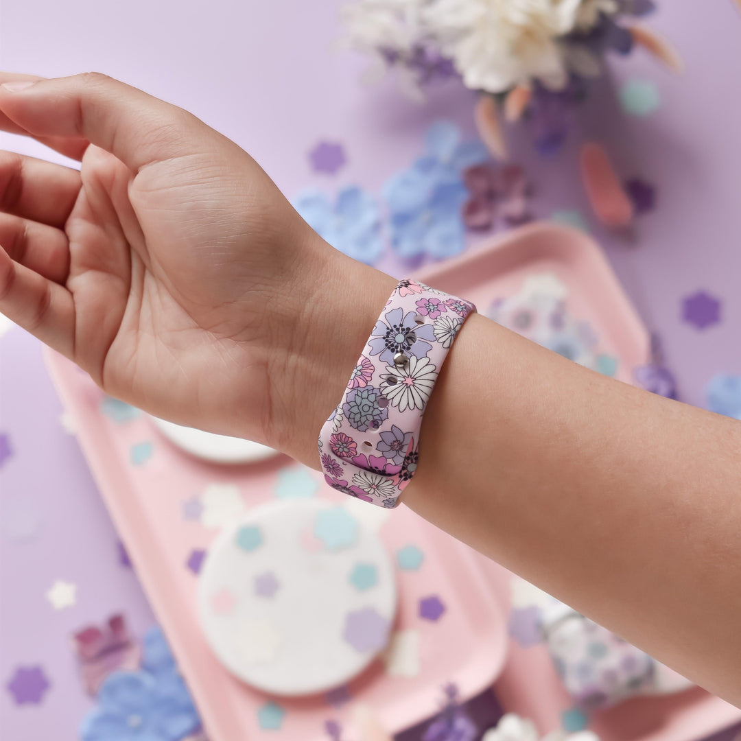 A hand is shown wearing the "Pastel Flowers Apple Watch Strap" by Coconut Lane, which features pink, purple, and white floral patterns. The background includes a light pink tray adorned with various pastel-colored, cut-out flower shapes scattered on it, set against a purple surface with additional flower decorations. Coordinating pastel shades and floral themes make this accessory perfect for matching phone cases or other straps for your Apple Watch.