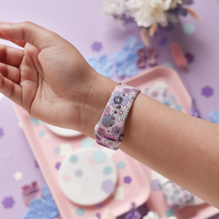 A hand is shown wearing the "Pastel Flowers Apple Watch Strap" by Coconut Lane, which features pink, purple, and white floral patterns. The background includes a light pink tray adorned with various pastel-colored, cut-out flower shapes scattered on it, set against a purple surface with additional flower decorations. Coordinating pastel shades and floral themes make this accessory perfect for matching phone cases or other straps for your Apple Watch.