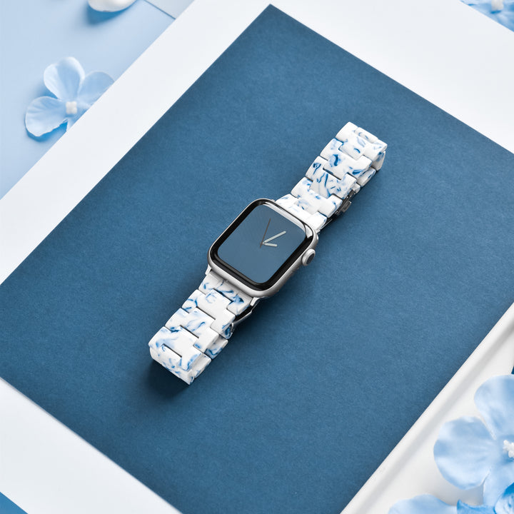A smartwatch featuring the Luxe Blue & White Apple Watch Strap by Coconut Lane is displayed on a dark blue rectangular background with white borders and decorative blue floral petals. The watch face is minimalistic, showcasing two hands over a solid blue background.
