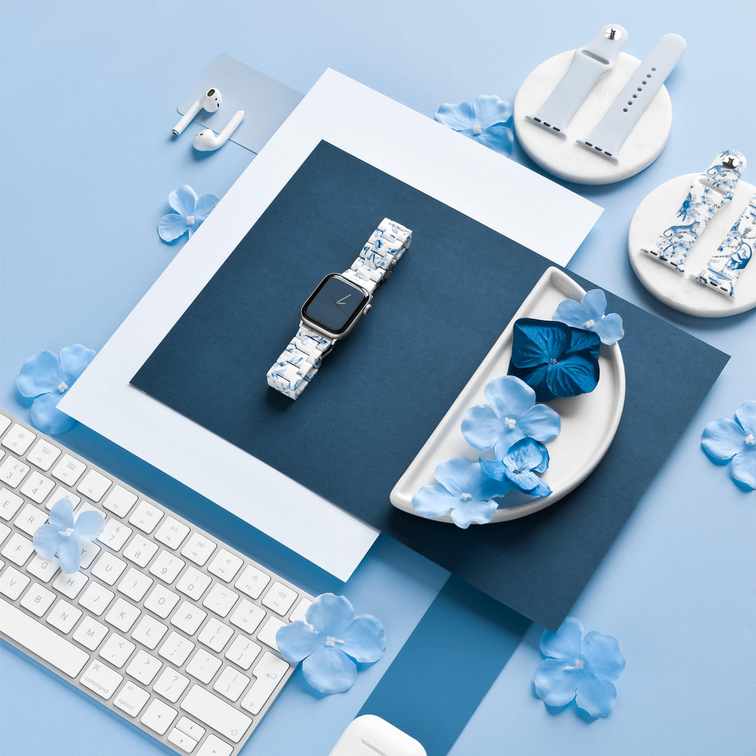 A flat lay photo featuring a Coconut Lane Luxe Blue & White Apple Watch Strap on blue and white paper backgrounds. Surrounding items include a keyboard, AirPods, ceramic holders, and blue flower petals arranged artistically. The color theme is predominantly blue and white.