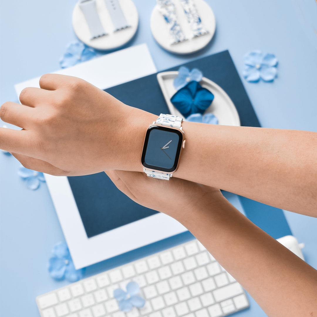 A person's wrist wearing a smartwatch with a Luxe Blue & White Apple Watch Strap by Coconut Lane is centered in the image. Surrounding the wrist are items in shades of blue and white, including paper, decorative flowers, and a keyboard, all arranged on a light blue surface.