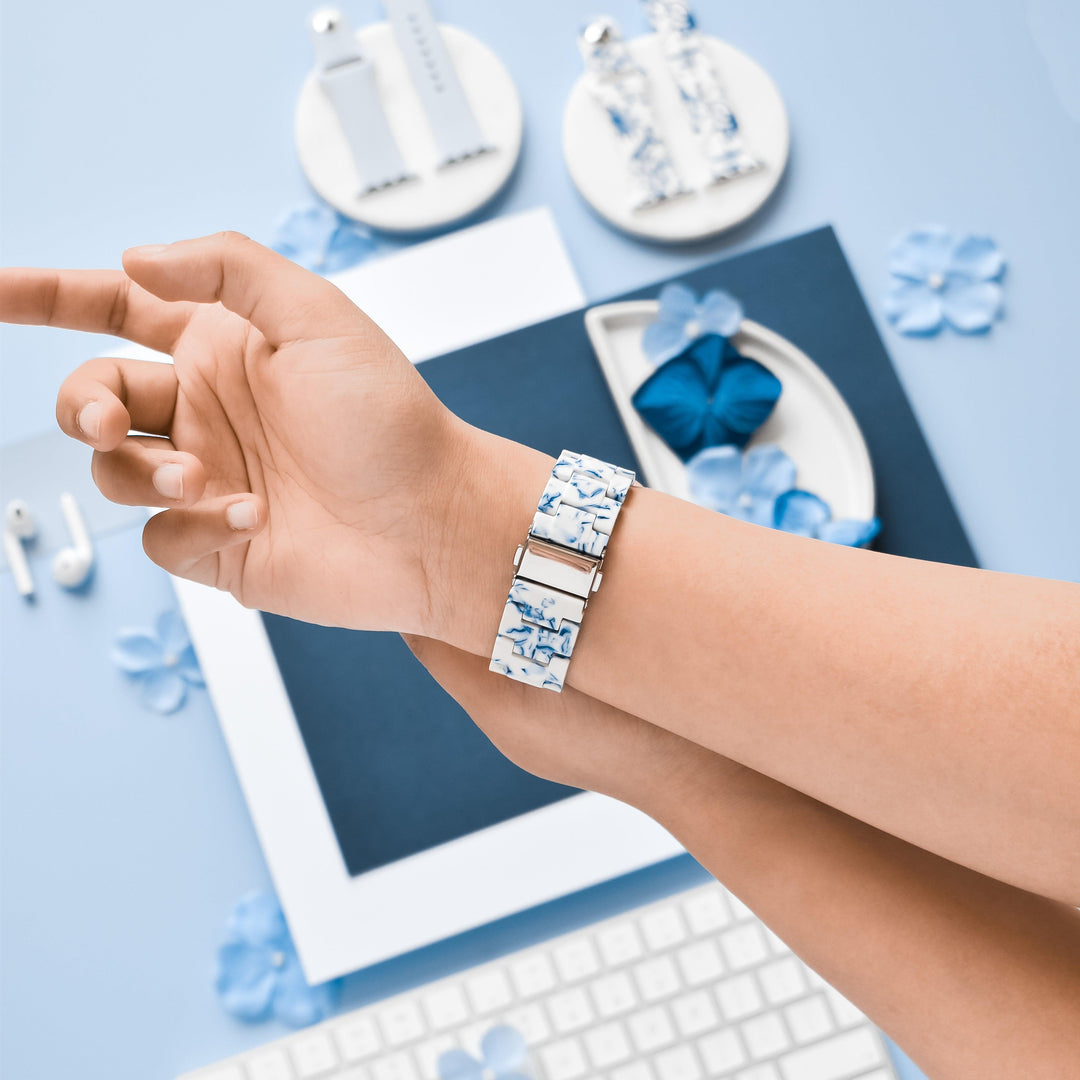A person wearing the Luxe Blue & White Apple Watch Strap by Coconut Lane points their finger. The background includes a white keyboard, floral decorations, a blue origami butterfly, earphones, and circular earring stands arranged on a light blue surface.