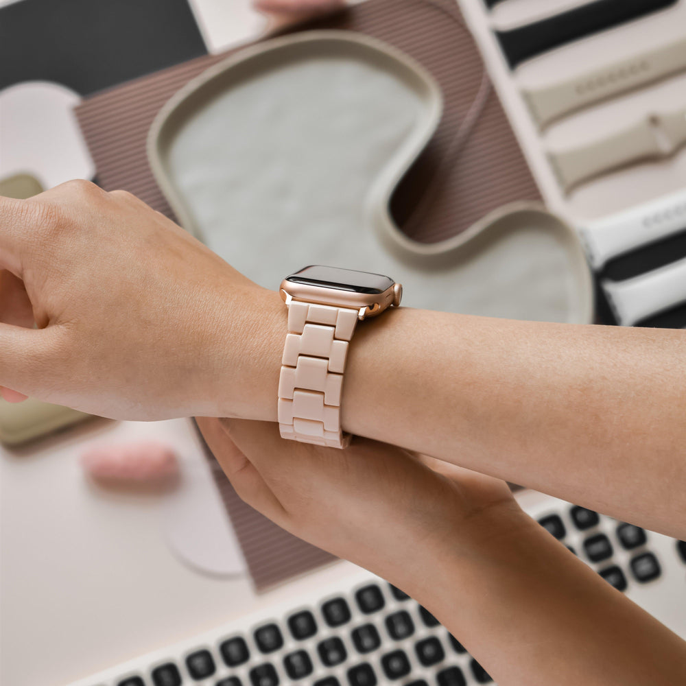 A person wears a rose gold smartwatch with a Luxe Nude Apple Watch Strap by Coconut Lane while resting their arm on a table. The table holds several smartwatch bands, including silicone, metal, and resin Apple Watch straps, as well as a partial view of a keyboard and a wavy-shaped tray.