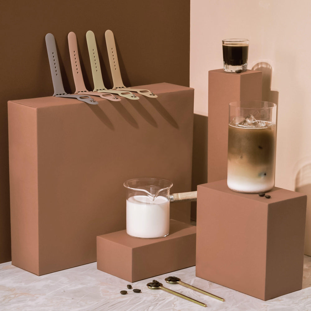 A minimalistic composition from the NAKD Collection featuring three brown boxes of varying heights. Displayed are four colored Coconut Lane Apple Watch Straps in Cappuccino, a small beaker of milk, a glass of iced coffee on a glass block, and a shorter glass of espresso. Two golden spoons and coffee beans adorn the marble surface.